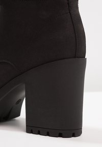 Bottine en daim noir avec un talon haut épais et une semelle texturée. Présente des éléments de design courbés et des coutures visibles le long des coutures.