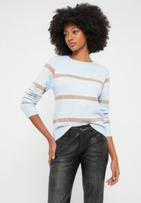 Moteris su garbanotais plaukais, dėvinti šviesiai mėlyną megztinį su kreminėmis juostelėmis ir tekstūruotas juodas odines kelnes, pozavanti viena ranka kišenėje.