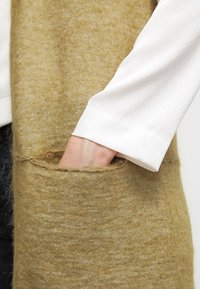 Lång, fluffig, senapsgul cardigan med synliga fickor. En vit, texturerad långärmad tröja är delvis synlig under.