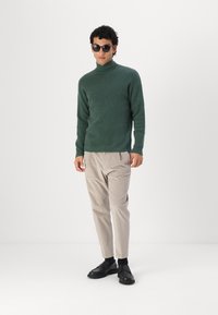 Zöld, bordázott garbós pulóver világos bézs, tapered nadrággal és fekete loaferekrel párosítva. A modellen kerek napszemüveg van. Minimalista háttér.
