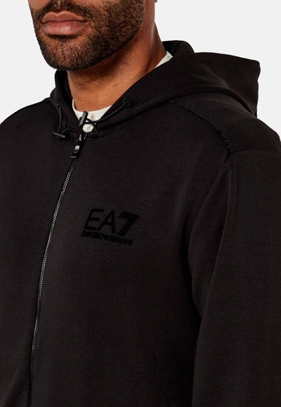Sudadera de cremallera negra hecha de tejido suave, con capucha ajustable y el logo de EA7 en el pecho, textura lisa y diseño entallado.