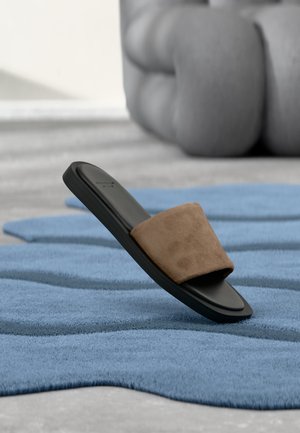 Zwarte sandaal met bruine suède band die rust op een gestructureerde blauwe mat met een groot glad grijs afgerond object op de achtergrond.