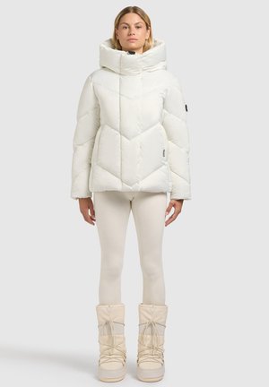 Witte gewatteerde jas met een hoge kraag, een chevron-gewatteerd ontwerp en twee zijzakken. Draagt bijpassende crème legging en sneeuwlaarzen.