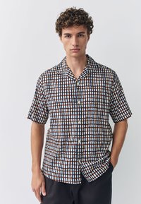 Camicia a maniche corte con motivo a griglia composta da linee blu, rosse e marroni su sfondo bianco. Presenta un colletto e una chiusura con bottoni sul davanti.