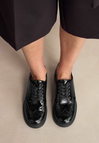Mustad nahast šnüreeritud kingad brogue detailidega, ümmarguste varvastega ja tekstureeritud viimistlusega, koos tumevärviliste lühikeste pükstega neutraalsel taustal.