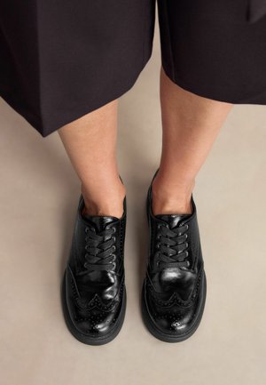 Czarne skórzane buty do wiązania z detalami w stylu brogue, okrągłe noski i teksturowane wykończenie, zestawione z ciemnymi spodnimi spodniami na neutralnym tle.