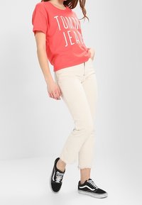 Korálové tričko s bílým textem "TOMMY JEANS", spárované s béžovými zkrácenými džíny a černými teniskami Vans s bílými tkaničkami.