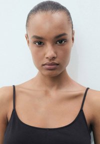 Top negro de tirantes finos con un diseño ajustado. La modelo tiene la piel suave y una expresión neutral, con el cabello recogido. Fondo liso.
