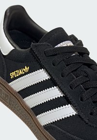Sapata de camurça preta com detalhes em couro branco e design de três riscas. Logotipo dourado "SPEZIAL" na lateral acima da sola de borracha textura.