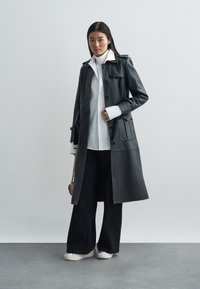Manteau trench en cuir noir avec un devant à boutons, des poches latérales et des épaulettes, associé à une chemise blanche et un pantalon noir à jambes larges.