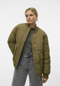 Olijf groene puffer jas met een geribde kraag, ritssluiting en voorzakken, gedragen over een zwart-wit gestreept shirt en grijze jeans.
