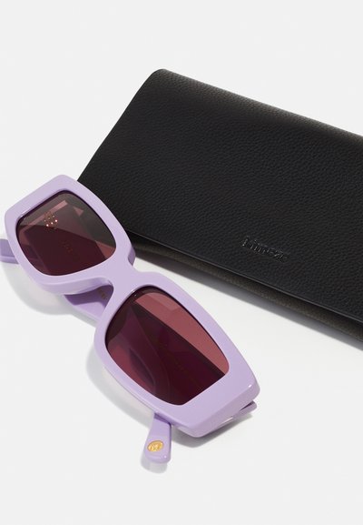 Lunettes de soleil violettes et surdimensionnées avec des montures angulaires et des verres rouge foncé, à côté d'une étui noir texturé embossé du nom de la marque.