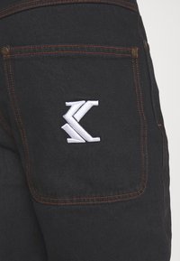 Svart denimjeans bakficka med orange sömnad och en vit broderad stiliserad "K"-logotyp centrerad på fickan.