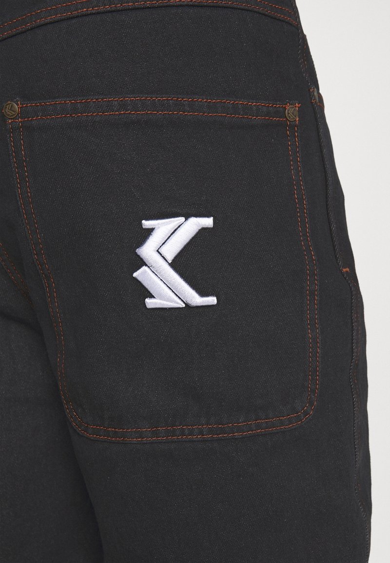 Svart denimjeans bakficka med orange sömnad och en vit broderad stiliserad "K"-logotyp centrerad på fickan.
