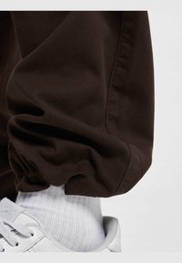 Bruine katoenen joggingbroek met elastische zoom, gecombineerd met een witte geribbelde sok en een witte sneaker met een textuur op het oppervlak.