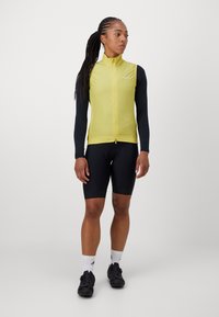 Damen-Radweste in leuchtendem Gelb mit hohem Kragen, Reißverschluss vorne und langen schwarzen Ärmeln. Dazu werden schwarze Shorts und Schuhe getragen.