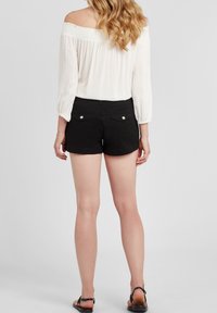Žena s dlouhými vlnitými vlasy, oblečená v bílé off-shoulder blůze, černých šortkách s zadními kapsami a černých sandálech, stojící zády.