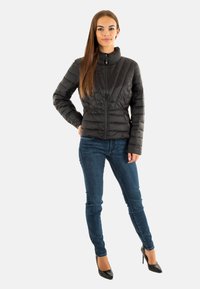 Giacca trapuntata nera con colletto alto, chiusura con zip e cuciture verticali, abbinata a jeans skinny blu e tacchi a spillo neri.