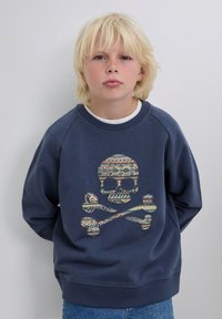 Niño rubio con ojos claros que lleva puesta una sudadera azul marino con un diseño de calavera y huesos cruzados, de pie contra un fondo sencillo.