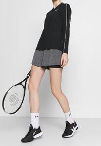 Zwart sportshirt met lange mouwen en witte zijstrepen, grijze geplooide shorts, zwarte sneakers en witte sokken. Houdt een tennisracket vast.