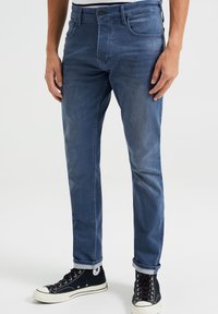 Ciemnoniebieskie dżinsy o wąskim kroju, z przednimi kieszeniami, szlufkami na pasek i zawiniętymi mankietami, w zestawie z czarnymi trampkami typu high-top.