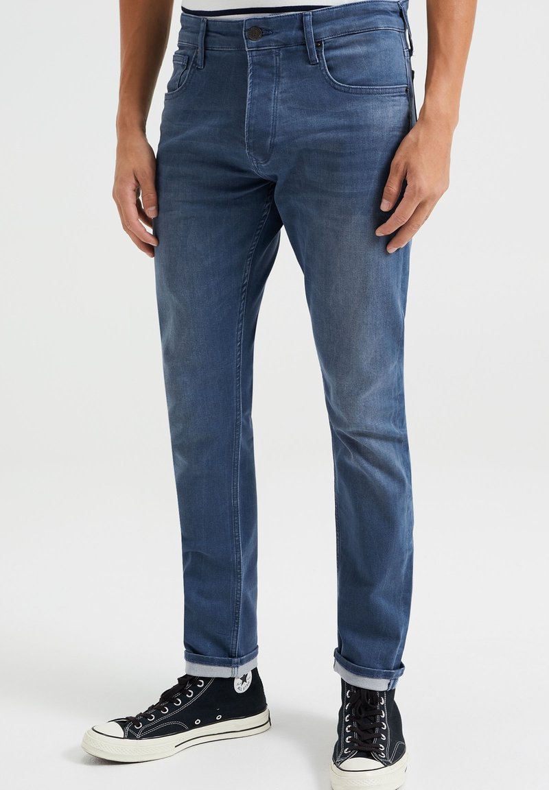 Ciemnoniebieskie dżinsy o wąskim kroju, z przednimi kieszeniami, szlufkami na pasek i zawiniętymi mankietami, w zestawie z czarnymi trampkami typu high-top.