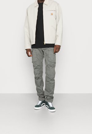 Veste gris clair avec une fermeture éclair à l'avant et un écusson logo, chemise noire, pantalon cargo gris avec plusieurs poches, et baskets montantes vertes.