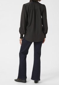 Chemise noir à manches longues avec des manches bouffantes et des poignets froncés, associée à un pantalon en denim foncé évasé et des chaussures plates noires.