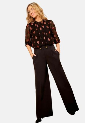 Blusa nera trasparente con motivi a cuori rossi, abbinata a pantaloni a vita alta neri con gamba ampia, dotati di bottoni dorati e tasche laterali.