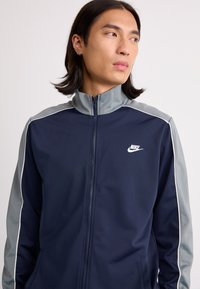 Navy zip-up jacket with gray accents, high collar, and white piping. Features a small white Nike logo on the chest. Smooth, athletic fabric.