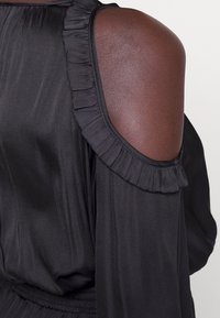 Svart plagg med en axelutskärning och volangdetaljer vid halslinningen. Slät textur med en subtil lyster. Samlad midja ger definition.