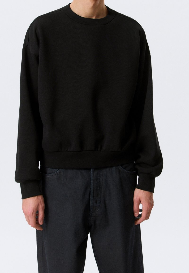 Personne portant un sweat-shirt noir uni à manches longues avec poignets et ourlet côtelés, associé à un pantalon foncé sur un fond clair.