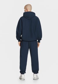 Granatowa bluza z kapturem o luźnym kroju, z prążkowanymi mankietami i pasującymi spodniami dresowymi. Materiał wydaje się miękki i teksturowany.