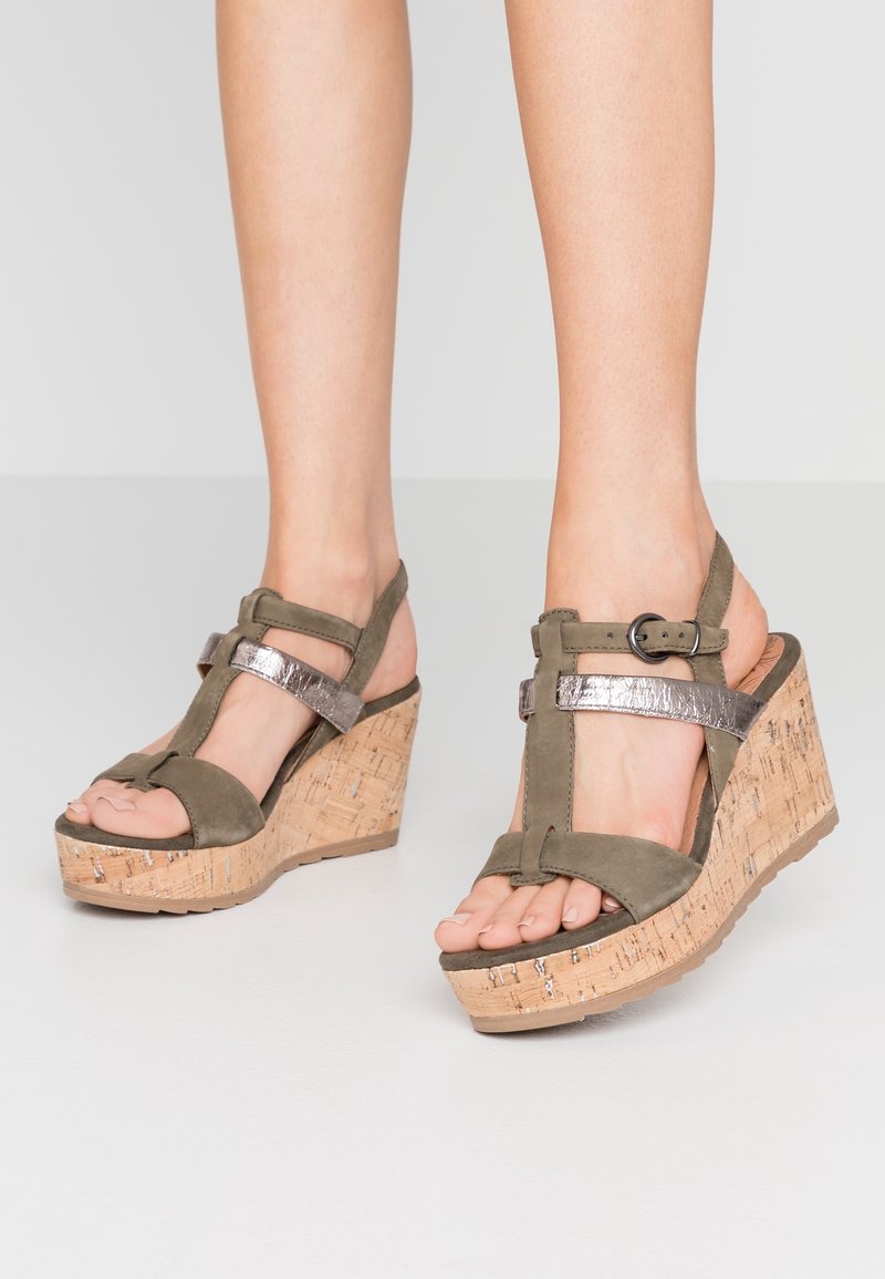 Sandales à talon compensé vert olive avec une plateforme en liège, des accents argent métalliques et une bride de cheville réglable. Design à bout ouvert avec une bride en T.