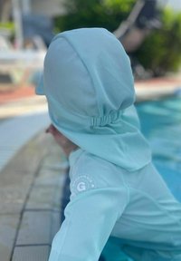 Vaaleansininen auringonsuojelupaidan uimapaita, jossa on huppu. Paidassa on tekstuuripinta ja ker gathered yksityiskohta kaulassa. Logo hihassa.