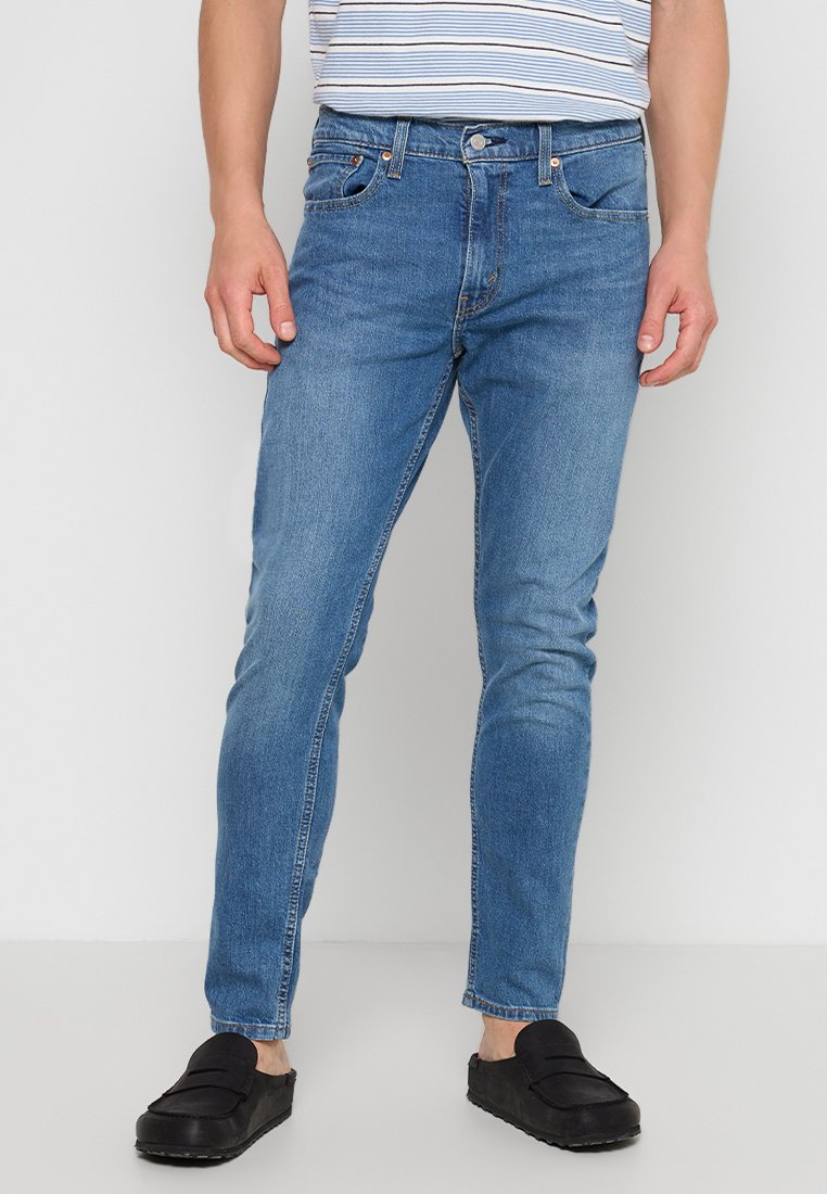 Mann trägt eine blaue Slim-Fit-Jeans, schwarze Slipper und ein weiß-blau gestreiftes Hemd, steht vor einem schlichten hellen Hintergrund.