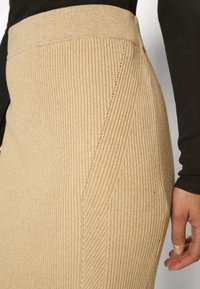 Beige žebrovaná pletená sukně s vysokým pasem, s diagonálními švy a texturovaným povrchem. Detailní záběr, ruka na pravé straně.