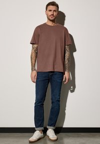 Hnědé tričko s kulatým výstřihem, volný střih, nošeno s tmavě modrými džíny a bílými teniskami. Tetování viditelné na pažích, stojící proti světlému pozadí.