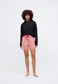 Zwarte hoodie met een rood logo, gecombineerd met roze shorts met een wit geometrisch patroon en een rode trekkoord.