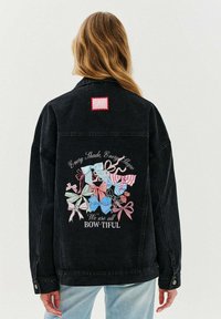 Czarna kurtka dżinsowa z kolorową grafiką różnych kokard na plecach, z napisem "Wszyscy jesteśmy BOW-TIFUL". Czerwona naszywka w kształcie kwadratu.