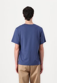 Navy blue cotton T-shirt with short sleeves and a rounded neckline, paired with khaki trousers. Back view, no visible patterns or accents.