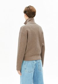 Bruine trui met hoge kraag gemaakt van een gestructureerde gebreide stof, gecombineerd met lichtblauwe jeans met wijde pijpen. Achteraanzicht.