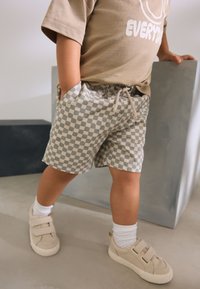 T-shirt beige et short à carreaux beige avec poches, associé à des baskets claires et des chaussettes blanches. Tissu lisse avec une coupe décontractée.