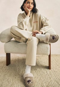 Beige loungewear with a zipper, fuzzy slippers featuring a teddy bear design, ribbed texture, and plush fur trim, sitting on a textured rug.
