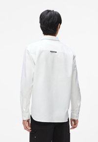 Ilga rankovė balta marškiniai, pagaminti iš medvilnės, su kreivos apykakle, sagomis ant rankovių ir juodu žymekliu nugaroje. Glotni tekstūra, klasikinis siluetas.