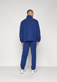 Σκούρος μπλε προπονητικός σκούφος με τζάκετ που κλείνει με φερμουάρ και παντελόνι που διαθέτει λευκές ρίγες στο πλάι. Το υλικό φαίνεται απαλό, με άνετη εφαρμογή και ελαστικές μανσέτες.