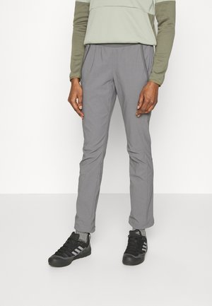 Person trägt graue Slim-Fit-Hose, schwarze Adidas-Sneaker und ein zweifarbiges Langarmshirt und steht vor einem schlichten Hintergrund.