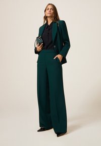 Traje verde a medida con pantalones de pierna ancha y una blazer ajustada. Camisa negra debajo, sosteniendo un pequeño bolso negro. Tela suave, diseño estructurado.