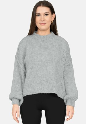 Eine Frau trägt einen locker sitzenden, hellgrauen Strickpullover mit Ballonärmeln und schwarze Hosen, steht mit dem Gesicht nach vorn vor einem einfarbigen Hintergrund.