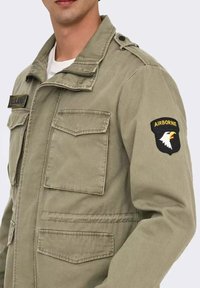 Giacca militare in khaki, misto cotone, con zip frontale, due tasche sul petto, un distintivo sulla spalla con un'aquila e dettagli con bottoni.